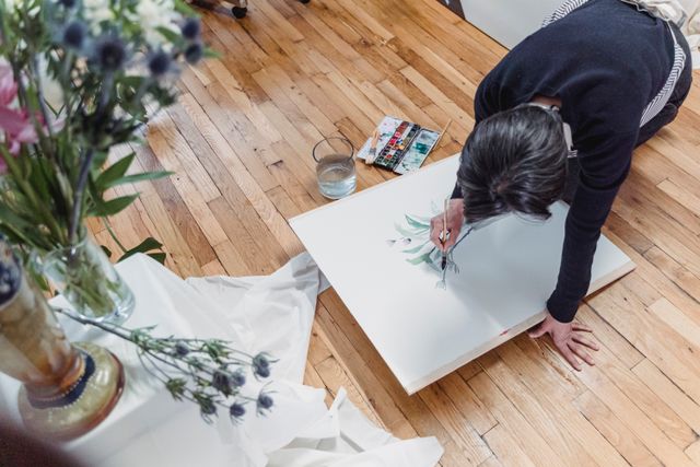 Eine Frau malt in einem hellen Atelier ein florales Motiv auf eine Leinwand.