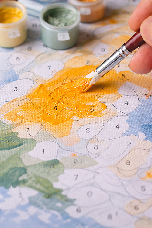 Nahaufnahme: Pinsel trägt gelb-orange Acrylfarbe auf eine Malen-nach-Zahlen-Vorlage auf; die Fläche wirkt noch ungleichmäßig, Farbtöpfchen stehen im Hintergrund.