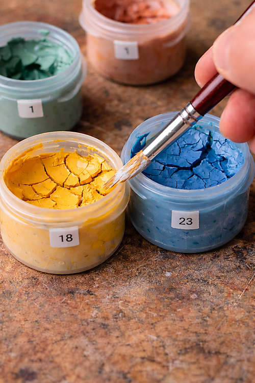 Nahaufnahme von geöffneten Farbtöpfchen mit rissig eingetrockneter Acrylfarbe; ein Pinsel berührt die Oberfläche – typisches Problem bei Malen-nach-Zahlen-Sets.