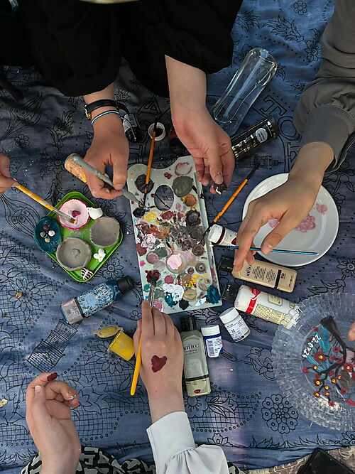 Nahaufnahme von Händen beim Malen mit Pinsel auf einer Leinwand; Farben und Malutensilien liegen bereit – Symbolbild für gemeinsames Malen nach Zahlen zu zweit oder in der Familie.