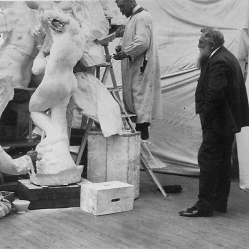 Schwarzweißfoto eines Bildhauerateliers: Mehrere Männer in hellen Arbeitskitteln bearbeiten eine große Gipsgruppe, während Auguste Rodin rechts im dunklen Anzug zusieht.