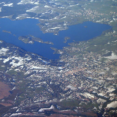 Weiter Blick über den Staffelsee bei Murnau mit bewaldeten Ufern, kleinen Inseln und einer hügeligen Landschaft unter bewölktem Himmel.