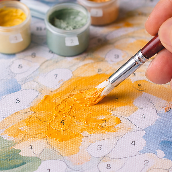 Nahaufnahme: Pinsel trägt gelb-orange Acrylfarbe auf eine Malen-nach-Zahlen-Vorlage auf; die Fläche wirkt noch ungleichmäßig, Farbtöpfchen stehen im Hintergrund.