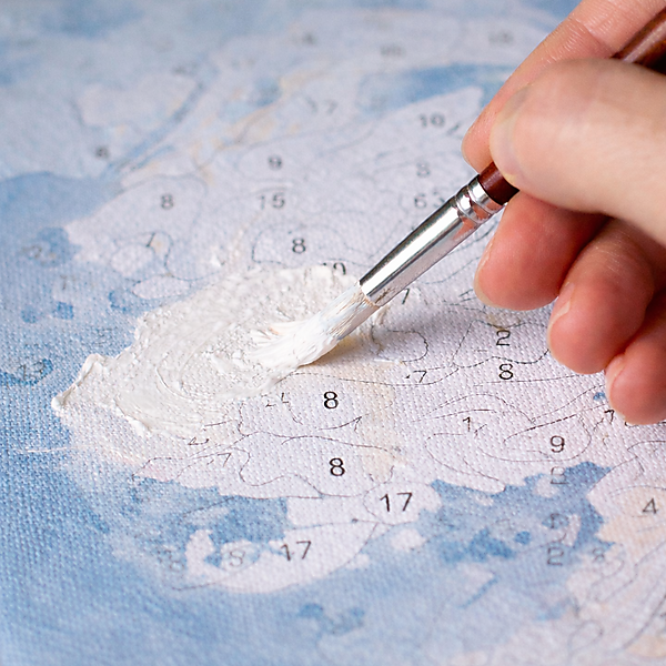 Hand trägt weiße Acrylfarbe mit einem Pinsel auf eine Malen-nach-Zahlen-Vorlage auf, um aufgedruckte Zahlen zu überdecken.