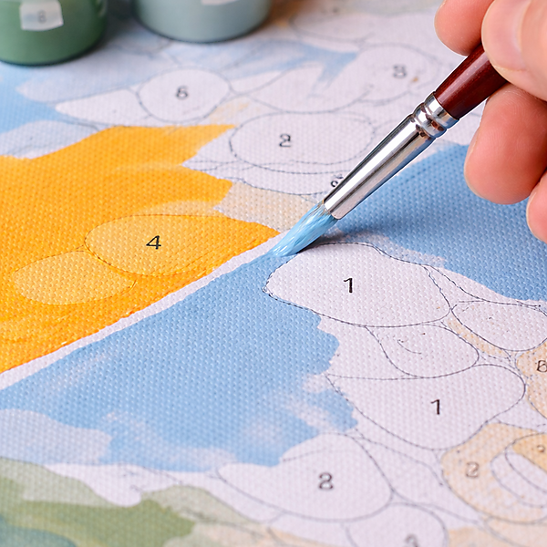 Nahaufnahme: Hand zieht mit einem feinen Pinsel eine saubere Kante zwischen blauer und gelb-oranger Fläche auf einer Malen-nach-Zahlen-Leinwand; Farbtöpfe sind im Hintergrund zu sehen.