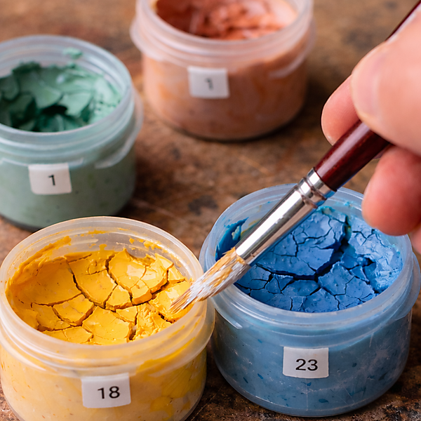 Nahaufnahme von geöffneten Farbtöpfchen mit rissig eingetrockneter Acrylfarbe; ein Pinsel berührt die Oberfläche – typisches Problem bei Malen-nach-Zahlen-Sets.