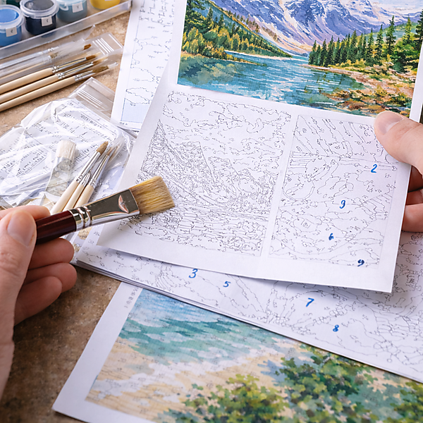 Hände halten eine Malen-nach-Zahlen-Vorlage; Farbtöpfe, Pinsel und ein Farbfächer liegen auf einem Tisch – Symbolbild für Kaufberatung und Materialauswahl.