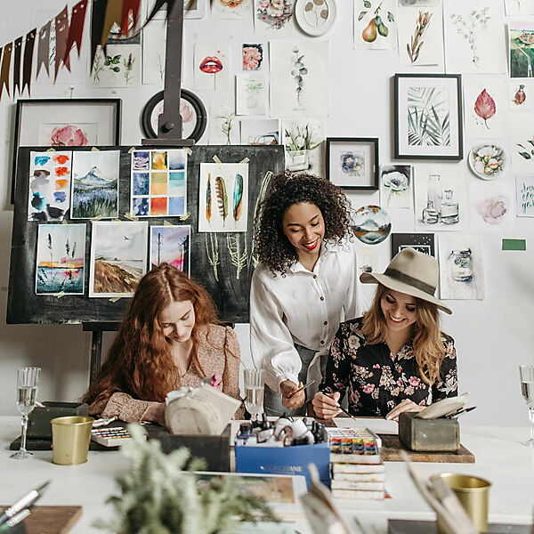 Mehrere Menschen sitzen an einem Tisch und malen gemeinsam; Pinsel, Farben und Blumen stehen bereit – Symbolbild für einen entspannten Malabend (Paint-Night) zu Hause.