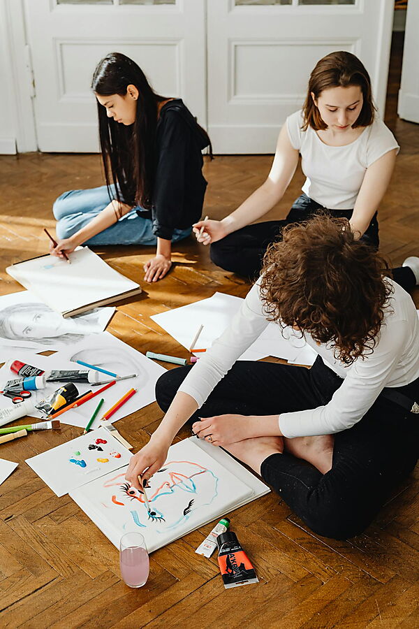 Mehrere Frauen sitzen gemeinsam auf dem Boden in einer kreativen Arbeitssituation und beschäftigen sich mit Zeichnen und Malerei.
