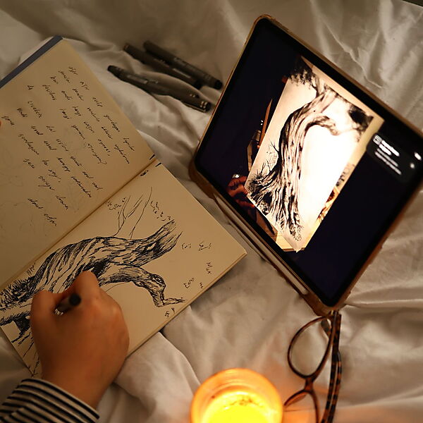 Hand zeichnet einen Baum in ein Skizzenbuch und nutzt dabei eine Vorlage auf dem Tablet.
