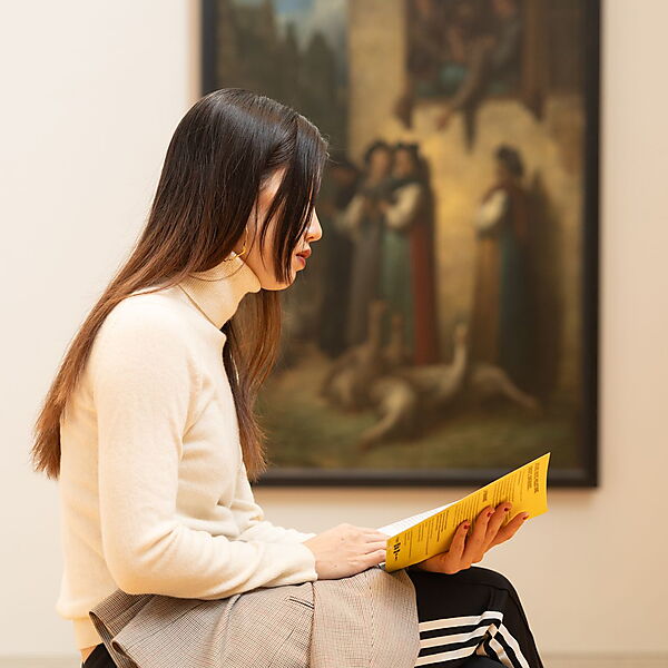 Frau sitzt in einem Museum auf einer Bank und liest eine Broschüre, um sich einem Kunstwerk Schritt für Schritt zu nähern.