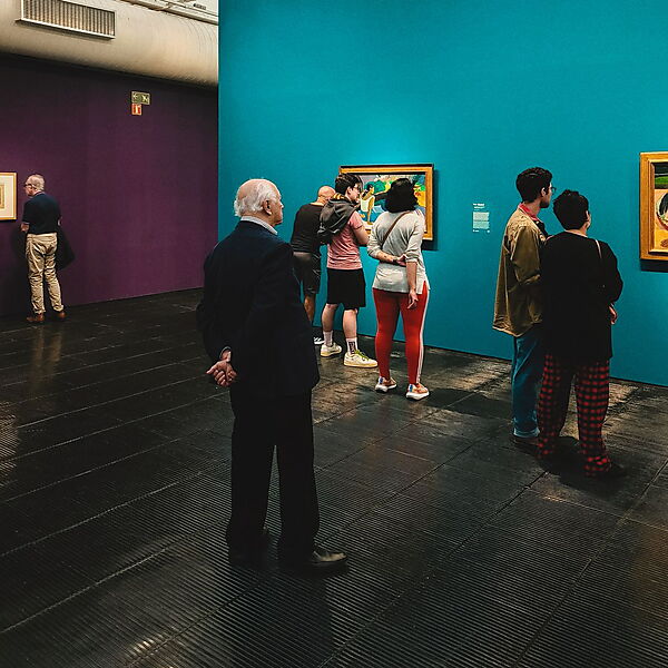 Besucher betrachten Gemälde in einer Galerie mit farbigen Wänden, die die Wirkung der ausgestellten Bilder deutlich mitprägen.