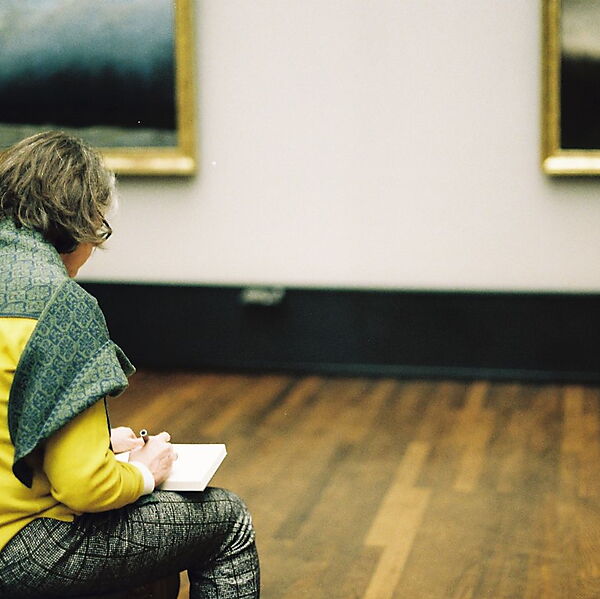 Person sitzt in einer Kunstgalerie und skizziert vor ausgestellten Bildern, um den eigenen Blick beim Zeichnen zu schulen.