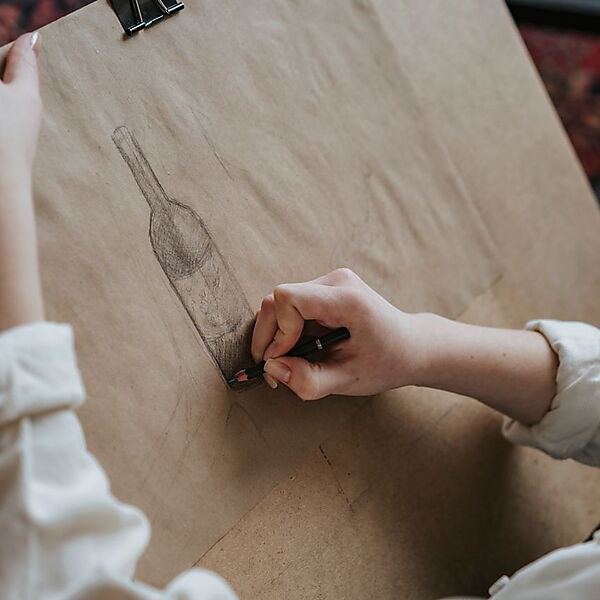 Eine Hand schattiert mit dem Bleistift die Zeichnung einer Flasche auf braunem Papier.