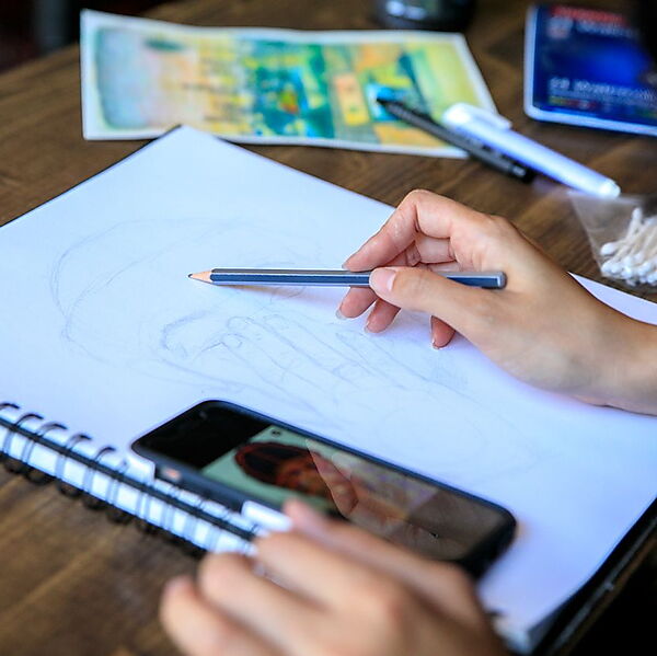Person zeichnet mit Bleistift in ein Skizzenbuch und nutzt ein Smartphone als Referenz für die entstehende Zeichnung.