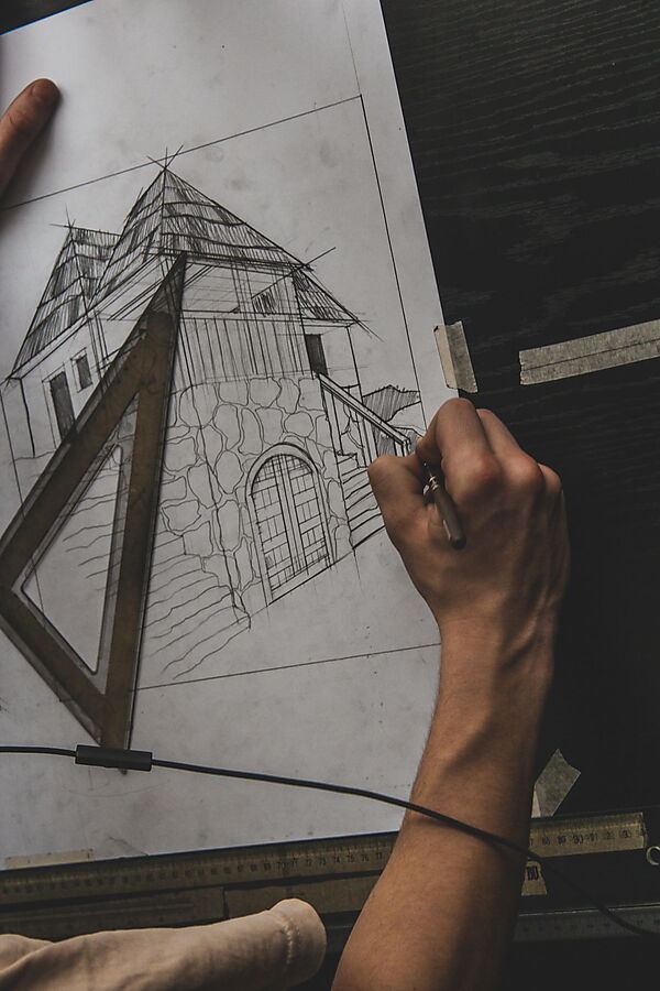 Hand zeichnet mit Bleistift und Lineal ein Haus auf Papier und prüft dabei Linien, Winkel und Perspektive.