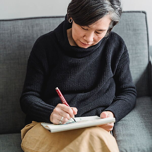 Erwachsene Frau sitzt entspannt auf einem Sofa und zeichnet konzentriert in ein Skizzenbuch.