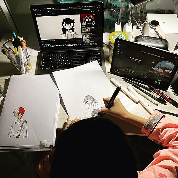 Eine Person zeichnet Figuren auf Papier, während auf einem Laptop ein Zeichentutorial läuft.