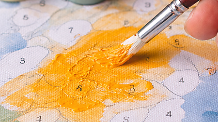 Nahaufnahme: Pinsel trägt gelb-orange Acrylfarbe auf eine Malen-nach-Zahlen-Vorlage auf; die Fläche wirkt noch ungleichmäßig, Farbtöpfchen stehen im Hintergrund.