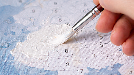 Hand trägt weiße Acrylfarbe mit einem Pinsel auf eine Malen-nach-Zahlen-Vorlage auf, um aufgedruckte Zahlen zu überdecken.