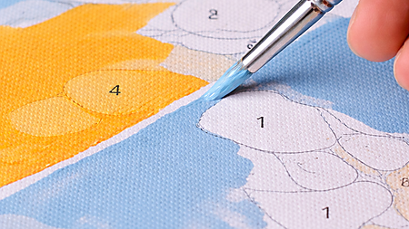 Nahaufnahme: Hand zieht mit einem feinen Pinsel eine saubere Kante zwischen blauer und gelb-oranger Fläche auf einer Malen-nach-Zahlen-Leinwand; Farbtöpfe sind im Hintergrund zu sehen.