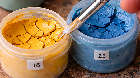 Nahaufnahme von geöffneten Farbtöpfchen mit rissig eingetrockneter Acrylfarbe; ein Pinsel berührt die Oberfläche – typisches Problem bei Malen-nach-Zahlen-Sets.