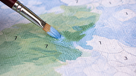 Nahaufnahme: Pinsel korrigiert einen Farbrand auf einer Malen-nach-Zahlen-Vorlage, indem hellblaue Farbe über eine kleine grüne Fehlstelle gestrichen wird; Farbtöpfchen sind im Hintergrund unscharf.