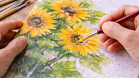 Nahaufnahme: Hände malen mit einem feinen Pinsel ein Sonnenblumen-Motiv auf einer Malen-nach-Zahlen-Leinwand; Farbtöpfchen und Pinsel liegen auf dem Tisch.