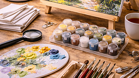 Arbeitsplatz mit Malen-nach-Zahlen-Set auf einer Staffelei, vielen Farbtöpfchen, Pinselset, Mischpalette, Lupe und heller Lampe – Übersicht über sinnvolles Zubehör.