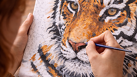 Nahaufnahme: Hände malen ein Malen-nach-Zahlen-Tigermotiv; nummerierte Flächen und Farbtöpfchen liegen bereit – Symbolbild für geführte Kreativität und ruhigen Rhythmus.