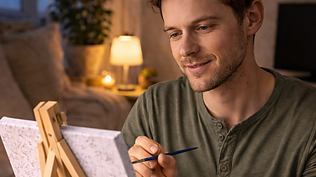 Junger Mann malt abends konzentriert an einer Malen-nach-Zahlen-Leinwand auf einer kleinen Staffelei; farbige Farbtöpfchen stehen vor der Staffelei auf dem Tisch, warmes Lampenlicht im Hintergrund.