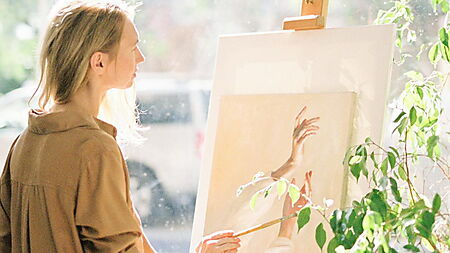 Frau steht nachdenklich neben einer grünen Zimmerpflanze – Symbolbild für Pause, Selbstfreundlichkeit und Neustart, wenn die Motivation beim Malen nach Zahlen fehlt.