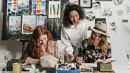 Mehrere Menschen sitzen an einem Tisch und malen gemeinsam; Pinsel, Farben und Blumen stehen bereit – Symbolbild für einen entspannten Malabend (Paint-Night) zu Hause.