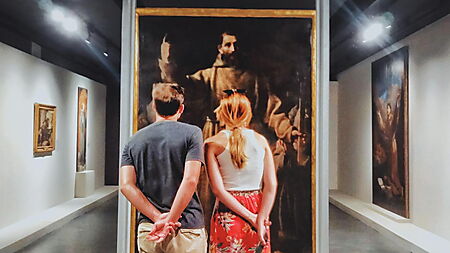 Ein im warmen Spotlicht gezeigtes Gemälde mit nächtlicher Straßenszene hängt in einer dunklen Galerie und lenkt den Blick auf Farbe, Licht und Komposition.