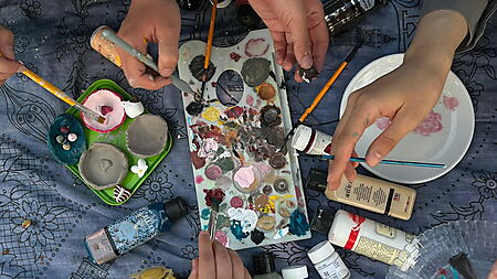 Nahaufnahme von Händen beim Malen mit Pinsel auf einer Leinwand; Farben und Malutensilien liegen bereit – Symbolbild für gemeinsames Malen nach Zahlen zu zweit oder in der Familie.