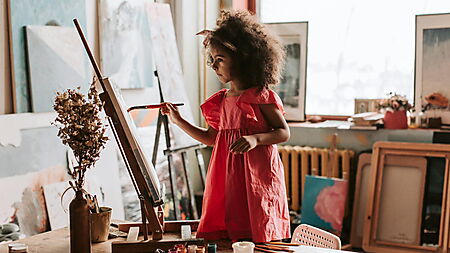 Kind malt an einer Leinwand; im Vordergrund stehen Blumen und Flaschen – Symbolbild für Maltechnik und sorgfältiges Arbeiten beim Malen nach Zahlen.