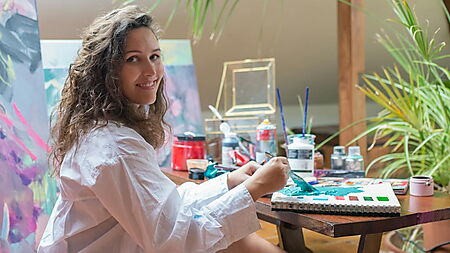 Frau arbeitet in entspannter Atelieratmosphäre mit Acrylfarben, Pinseln und Palette an einem Bild.