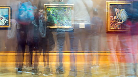 Verschwommene Besucher bewegen sich durch eine helle Galerie mit Gemälden und zeigen den Gegensatz von Bewegung und Ruhe im Bildraum.