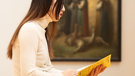 Frau sitzt in einem Museum auf einer Bank und liest eine Broschüre, um sich einem Kunstwerk Schritt für Schritt zu nähern.