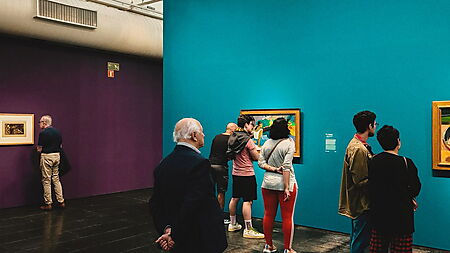 Besucher betrachten Gemälde in einer Galerie mit farbigen Wänden, die die Wirkung der ausgestellten Bilder deutlich mitprägen.