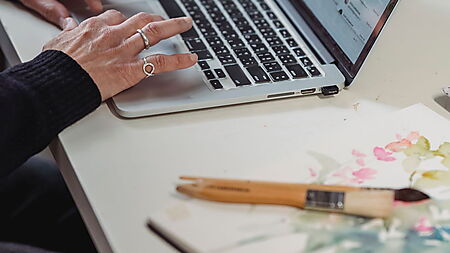 Frau zeichnet an einem Schreibtisch und nutzt Blumenbilder auf einem Laptop als Referenz für die eigene Zeichnung.