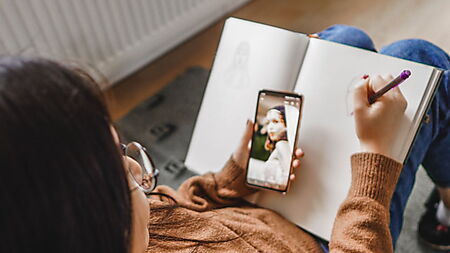 Frau zeichnet ein Porträt nach einer Smartphone-Vorlage und vergleicht dabei Vorstellung, Referenz und entstehende Zeichnung.