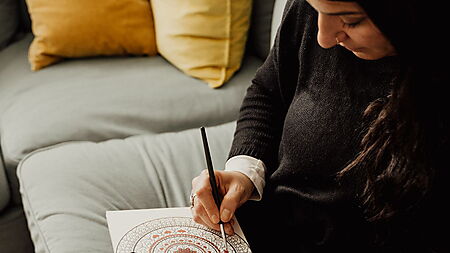 Frau malt entspannt ein farbenfrohes Mandala und macht daraus eine kleine kreative Auszeit.