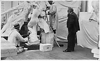 Schwarzweißfoto eines Bildhauerateliers: Mehrere Männer in hellen Arbeitskitteln bearbeiten eine große Gipsgruppe, während Auguste Rodin rechts im dunklen Anzug zusieht.
