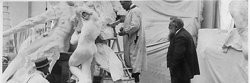Schwarzweißfoto eines Bildhauerateliers: Mehrere Männer in hellen Arbeitskitteln bearbeiten eine große Gipsgruppe, während Auguste Rodin rechts im dunklen Anzug zusieht.