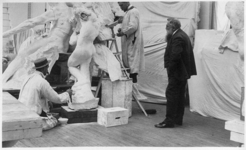 Schwarzweißfoto eines Bildhauerateliers: Mehrere Männer in hellen Arbeitskitteln bearbeiten eine große Gipsgruppe, während Auguste Rodin rechts im dunklen Anzug zusieht.