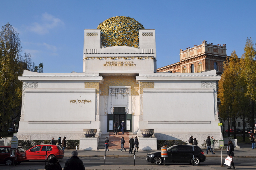Frontansicht des weißen Gebäudes der Wiener Secession mit symmetrischer Fassade, vergoldeter Lorbeerkuppel und dem Wahlspruch über dem Eingang.